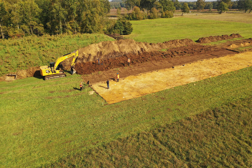Luchtfoto van een grasveld met daarachter bomen. Een deel van het grasveld is afgegraven door een gele graafmachine waardoor de aarde nu te zien is. Op het afgegraven gedeelte staat een vijftal mensen met oranje hesjes aan.