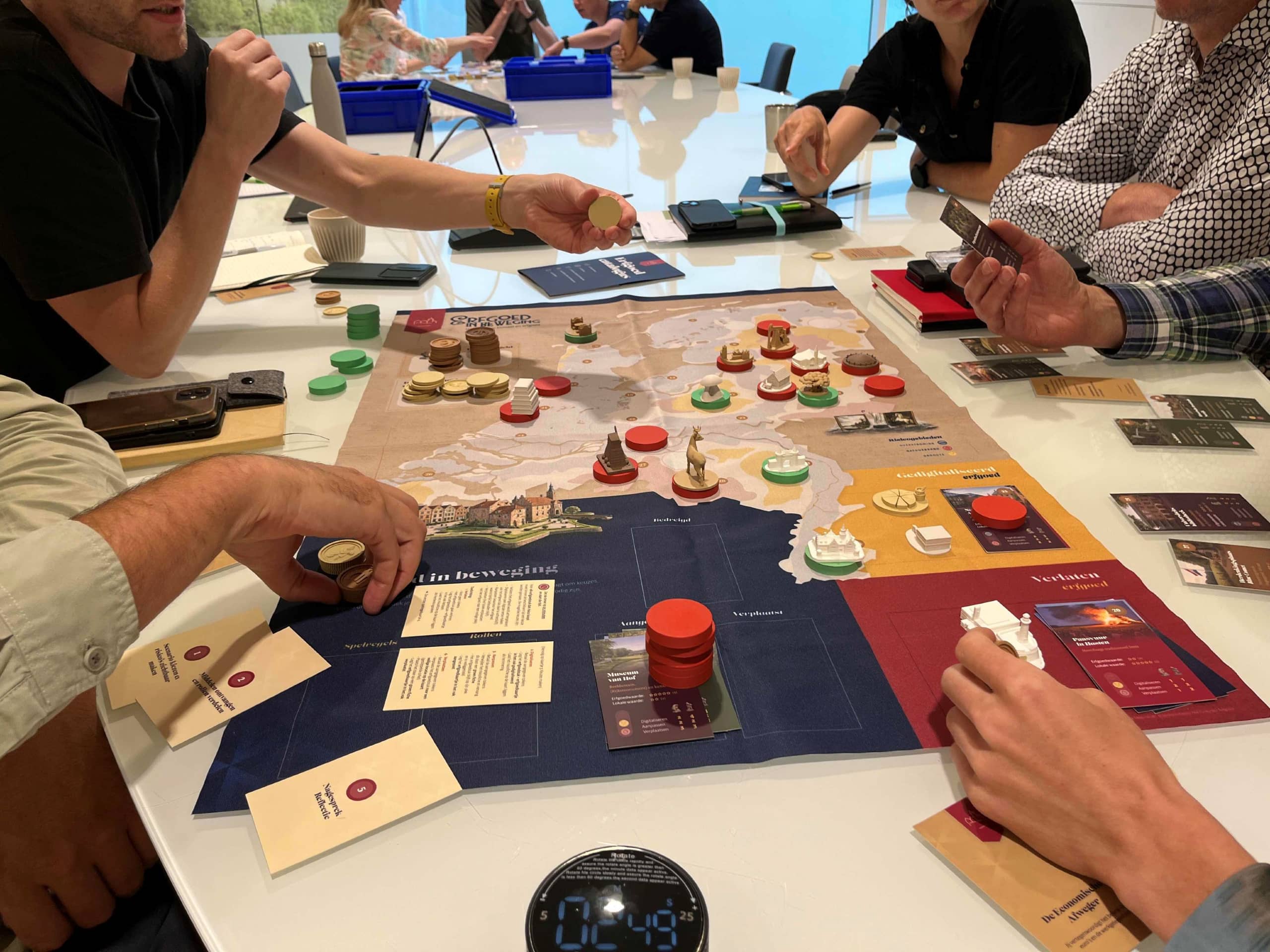 Vijf mensen, waarvan voornamelijk de handen zichtbaar zijn, spelen een bordspel aan een tafel in een zaal.