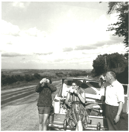 Een jongen, een vrouw en een man drinken en eten bij een auto langs de kant van de weg.