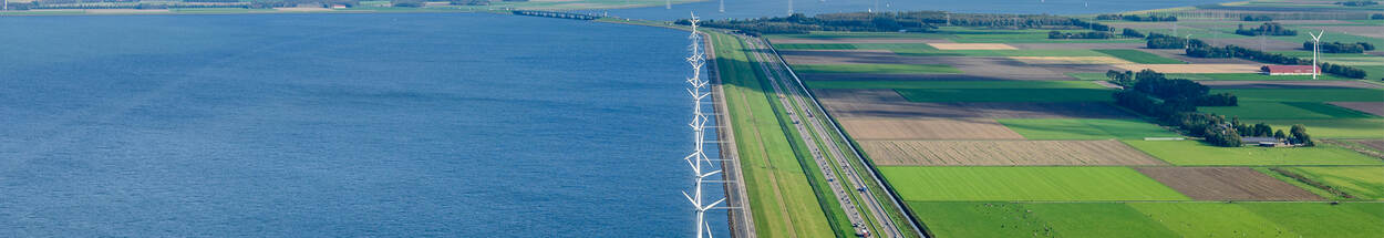 Luchtfoto van de IJsselmeerdijk met windmolens en de naastgelegen snelweg aan het water.