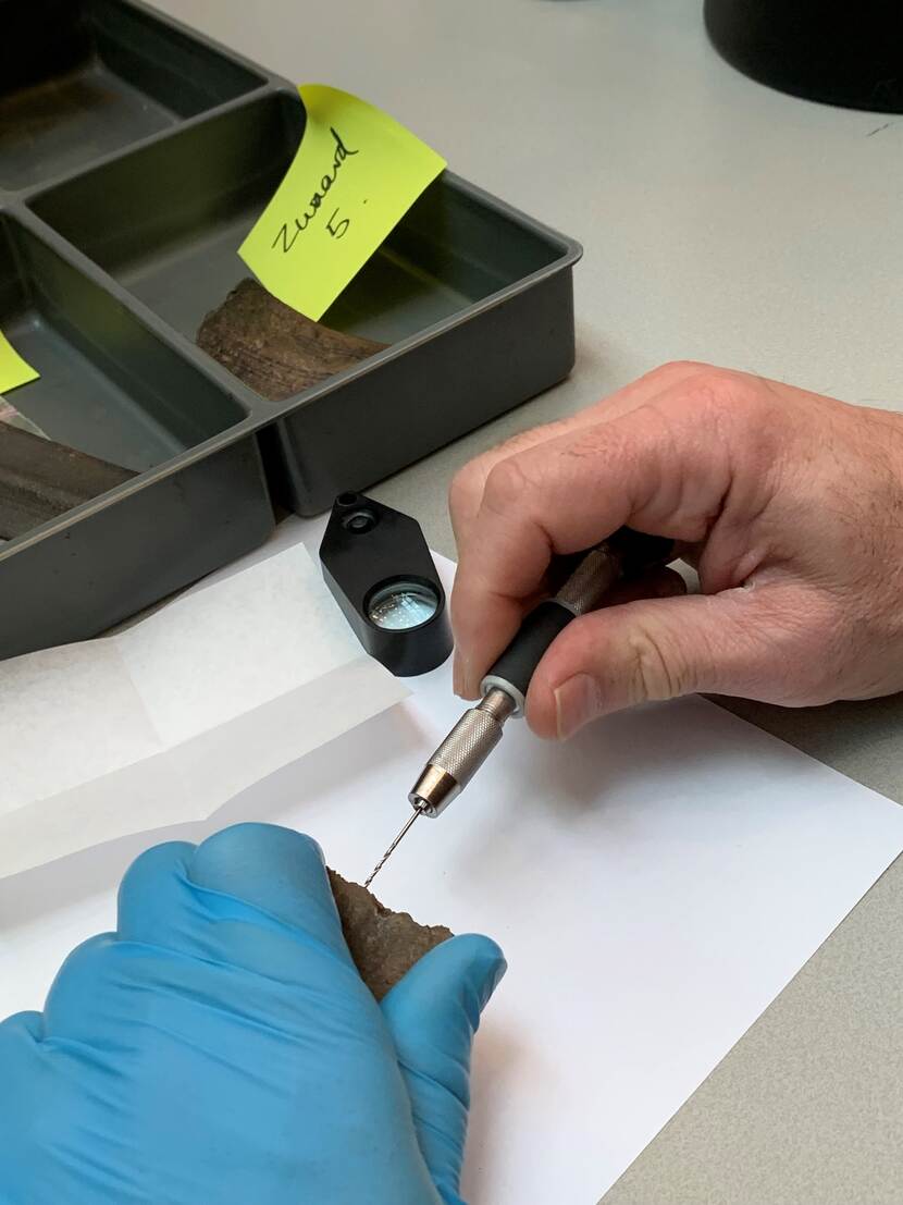 de hand van onderzoeker, die met een klein boortje een monster van een bronzen voorwerp neemt