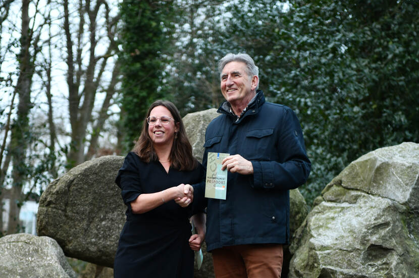 Gedeputeerde Jisse Otter (rechts) krijgt een archeologisch rijksmonumentenpaspoort overhandigd van Marjolein Verschuur van de RCE, met in de achtergrond een hunebed.