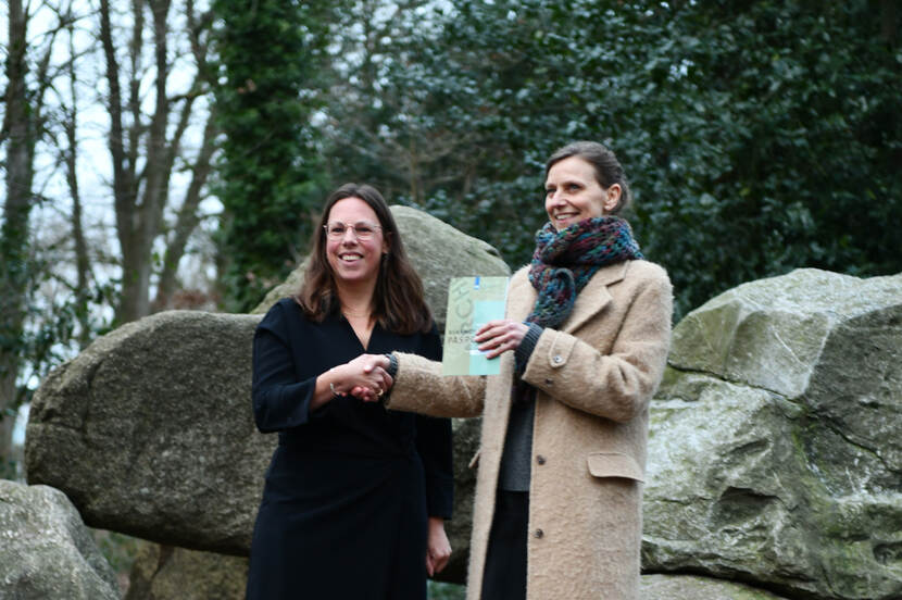 Sonja van der Meer (rechts) krijgt een archeologisch rijksmonumentenpaspoort overhandigd van Marjolein Verschuur van de RCE, met in de achtergrond een hunebed.