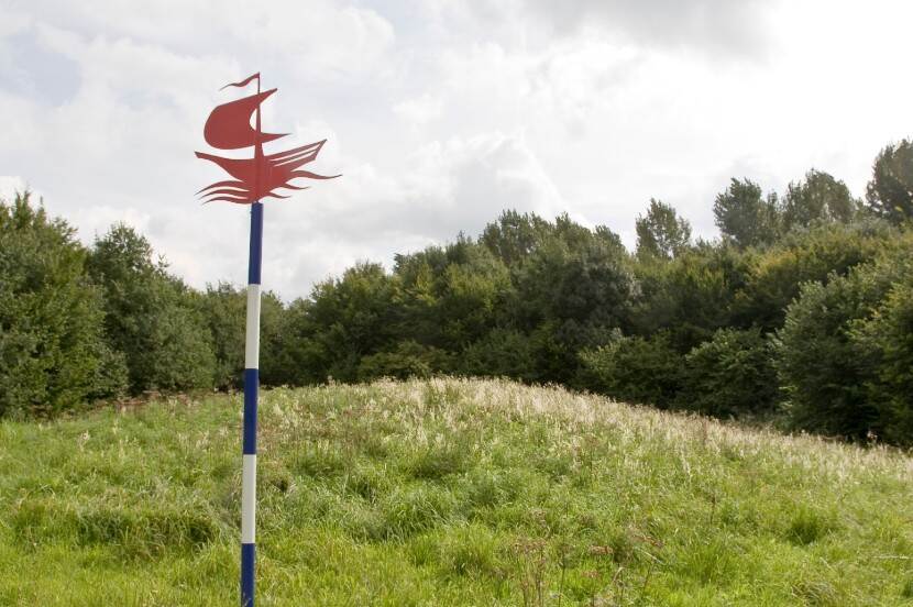 Een blauw wit paaltje met een rood scheepje daarop in het landschap markeert de plek waar een scheepswrak is gevonden.