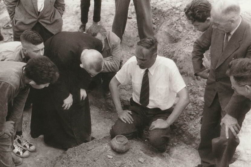 Prof. dr. Modderman, zittend te midden van vele toehoorders geeft aan de lokale geestelijke uitleg over de bandkeramische graven van Elsloo, 1966