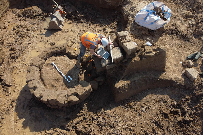 Luchtfoto van de opgraving van een waterput met een archeoloog en zijn laptop