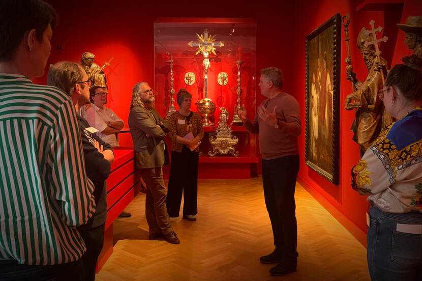 In een museumzaal met rode muren en verschillende religieuze voorwerpen staat een groep mensen rondom een spreker.