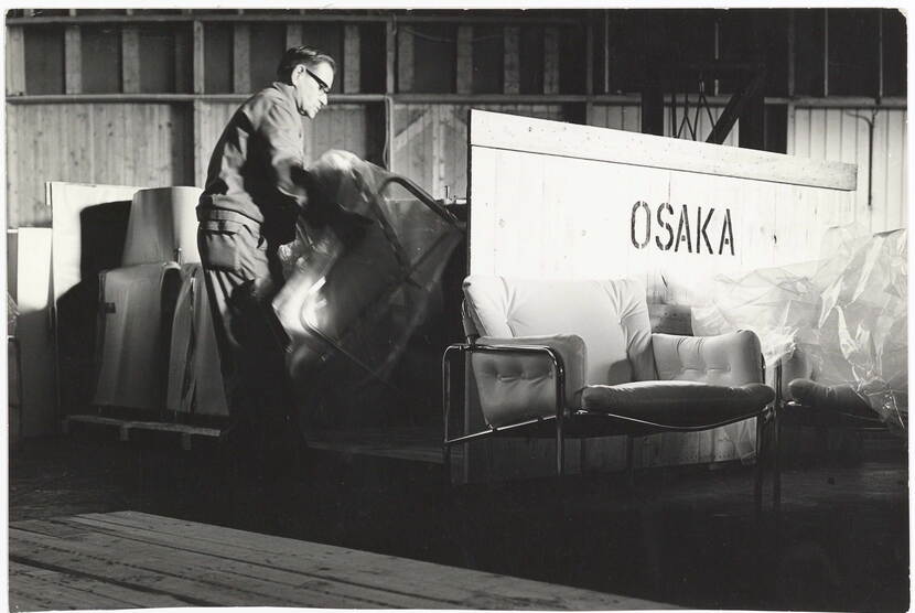 Zwart-wit foto van een man die een ingepakte stoel pakt uit een grote transportkrat waarop de naam Osaka op staat.