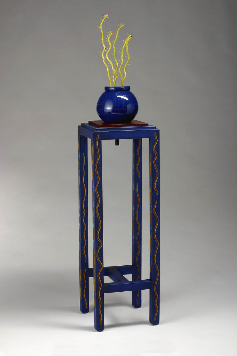 Hoog, blauw geschilderd, houten tafeltje, piedestal, met daarop een blauwe, glazen, bolvormige vaas met daarin goudgeel geschilderde, gestileerde, takken zonder bladeren.