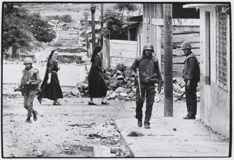 Foto van twee nonnen en drie gewapende  militairen in straat met vernielde gebouwen.