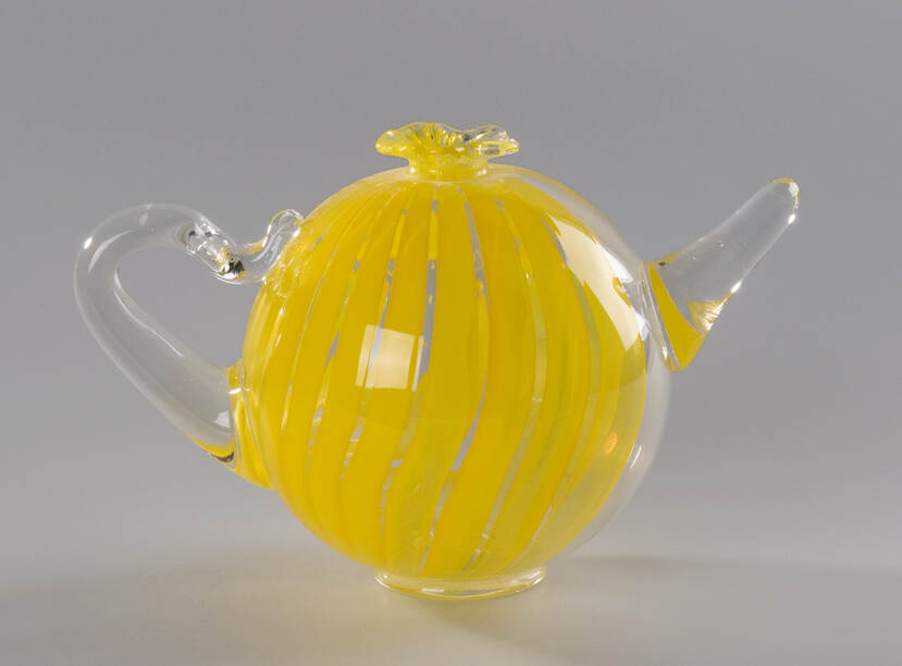 Glazen theepot versierd met gele banen. De tuit en het handvat zijn bevestigd aan de bolle vorm, en bovenaan zit een kleine uitwaaierende opening.