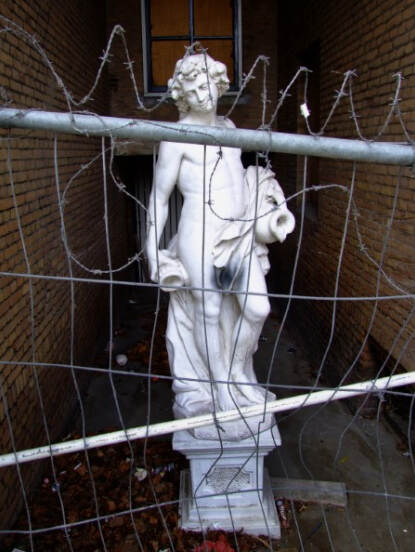 Een tuinbeeld is in een steeg geplaatst die is afgezet met een verbogen metalen afzettingshek en prikkeldraad waardoor deze ontoegankelijk is. Kunstenaar en datering onbekend.