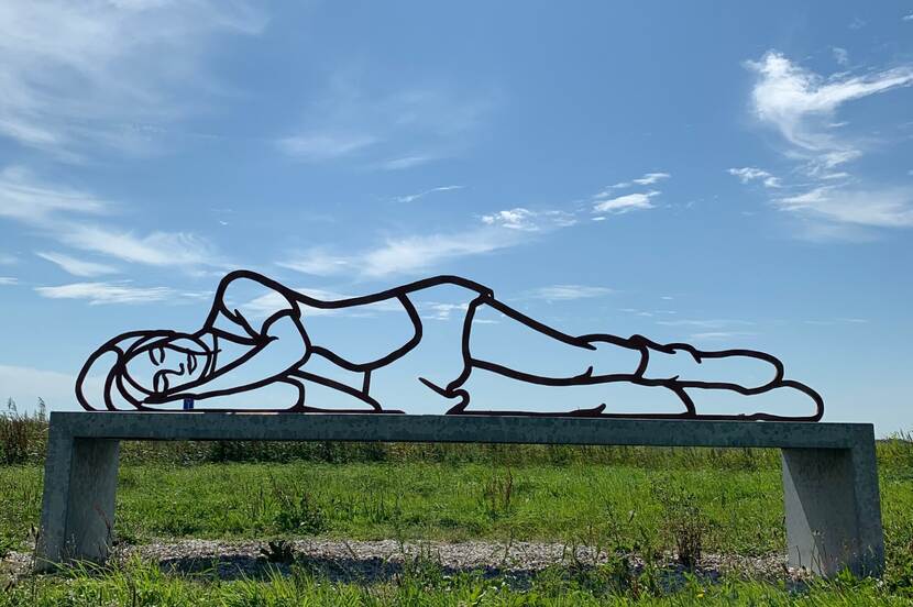 Beeld in de buitenlucht, genaamd Rustpunt van BlokLugthart uit 2014. Contouren van een grote liggende metalen mens-sculptuur die is neergestreken op een lange metalen bank, te midden in de natuur van het Groninger Hoogeland.
