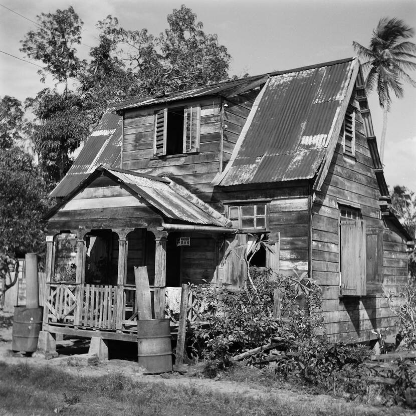 Zwartwit foto van een houten volkswoning in Albina, Suriname met lege olietonnen ervoor, golfplaten op het dak en palbomen in de achtergrond