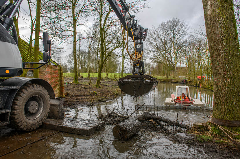 Een gracht omringd door bomen wordt gebaggerd door een machine.