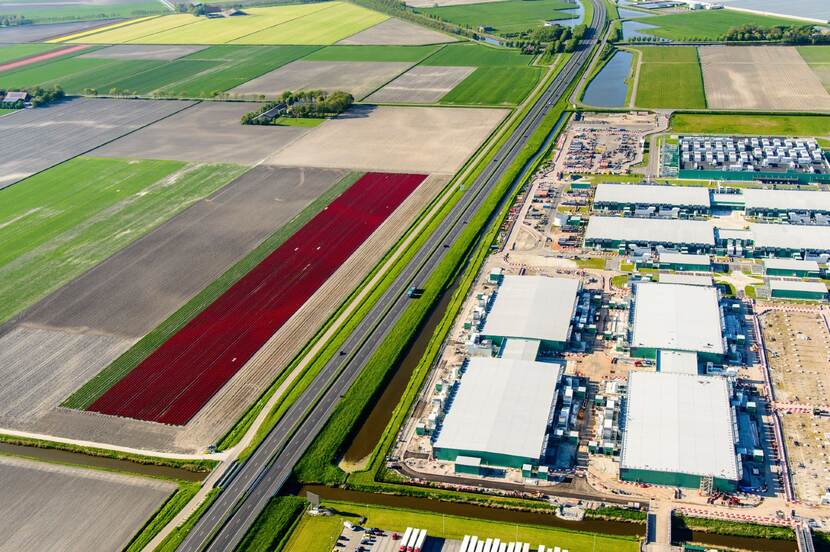 Foto van bovenaf een polder met links bloemenvelden en andere groene weilanden, dan een autoweg en daar rechts van grote rechthoekige gebouwen met datacentra erin.