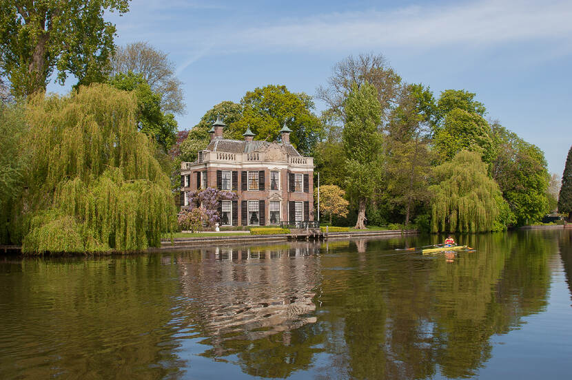 Roeier op een rivier met op de achtergrond een buitenplaats omringd door bomen en een helderblauwe lucht.