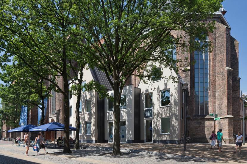 Kerkgebouw aan een zonnig plein in een stedelijke omgeving, met aan de voorzijde bomen met daaronder terrassen.