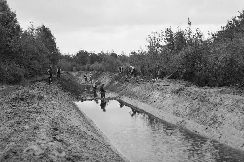 Foto van arbeiders die bezig zijn een beek uit te graven. Zichtbaar is de beek zelf, met zand en bomen en struiken eromheen.