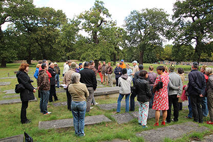 Een grote groep mensen tussen grafstenen die luisteren naar een verhaal. Op de achtergrond veel bomen