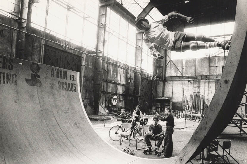 Zwart-wit foto van een jongen die op een halfpipe aan het skaten is, drie andere jongens zitten toe te kijken.