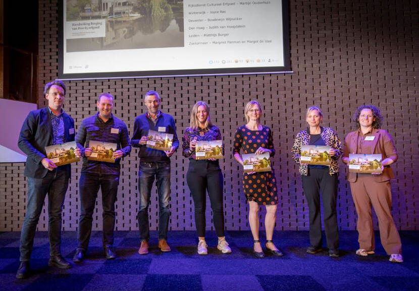 Groep van 8 mensen op het podium bij de uitreiking van het eerste exemplaar van de 'Handreiking borging' door Marijn Oosterhuis (RCE)