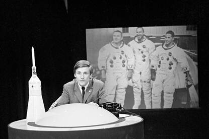 Henk Terlingen zit achter een ronde tafel met een ronde bolling in het midden, met op de achtergrond een afbeelding van astronauten.