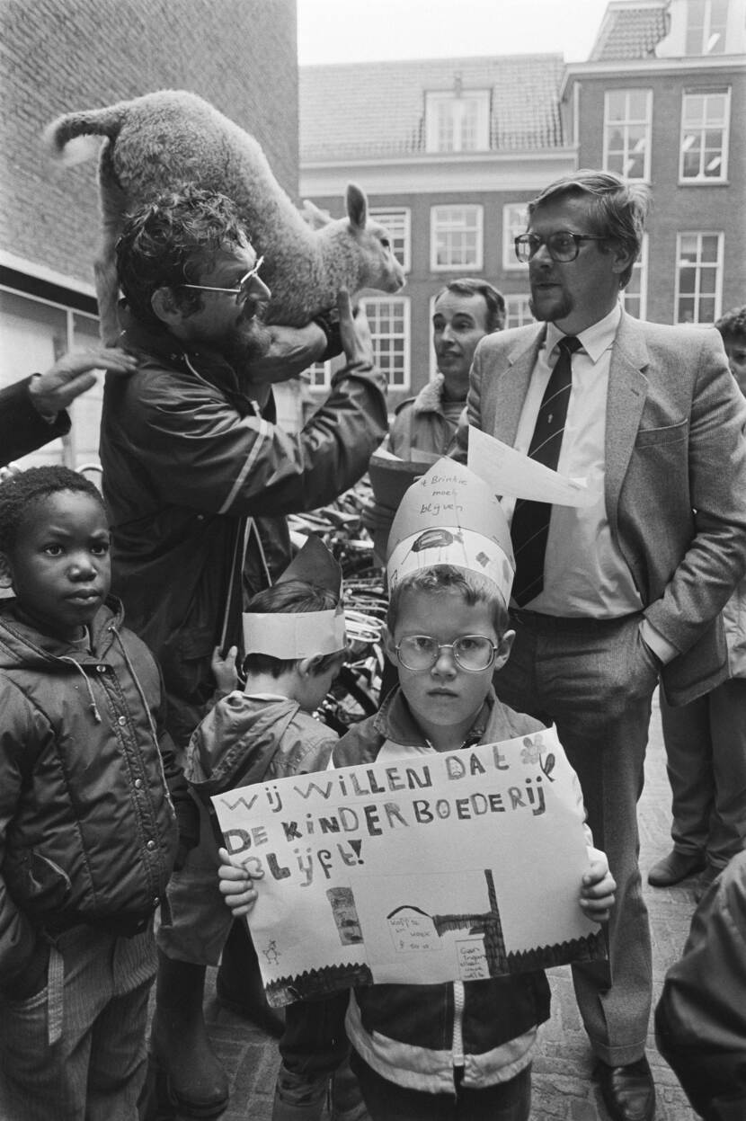 Het kind in het midden houd een papier vast met daarop "wij willen dat de kinderboederij blijft!". Verder staan er meer kinderen en volwassen mannen op de foto. Één man heeft een lam op zijn schouders terwijl hij praat met een andere man.