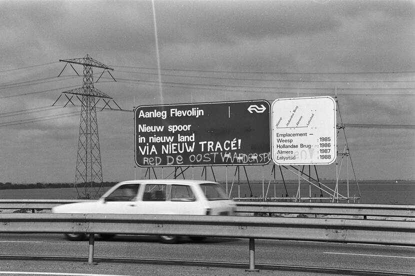 Zwart-wit foto van een auto op de snelweg die langs een bord rijdt waarbij de Flevolijn wordt aangekondigd. Daaronder is met een spuitbus geschreven: Red de Oostvaardsplassen!