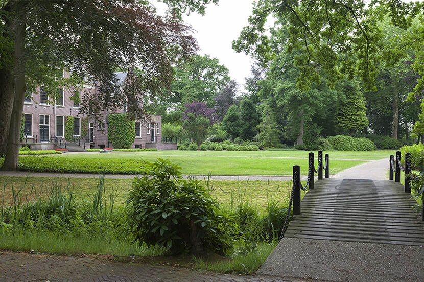 Park en tuinaanleg van Huis Hoevelaken met een houten bruggetje over een waterpartij en het huis half verscholen achter de grote parkbomen