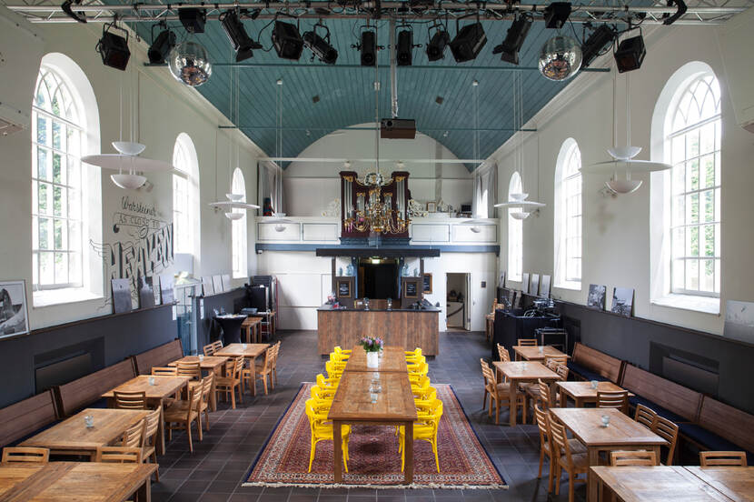 Interieur van een kerk in Borger die een nieuwe bestemming heeft gekregen. Zicht richting de witte achterwand met orgel. De kerk is ingericht als café met bar tafels en stoelen, met in het midden een kleed met daarop een lange tafel en acht knalgele stoelen.