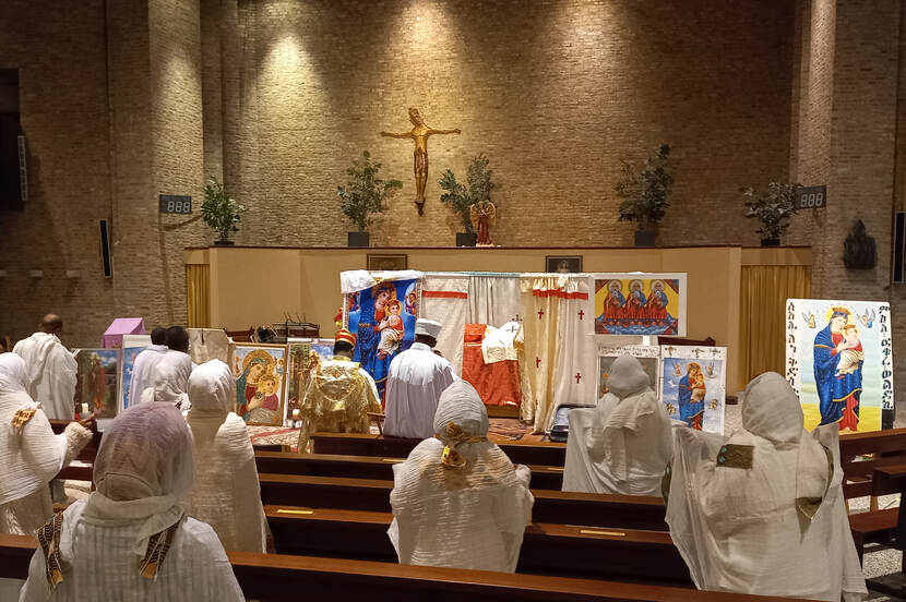 Interieur kerk met in witte gewaden geklede kerkgangers op de rug gezien, staand tussen de kerkbanken met hun gezichten richting altaar.