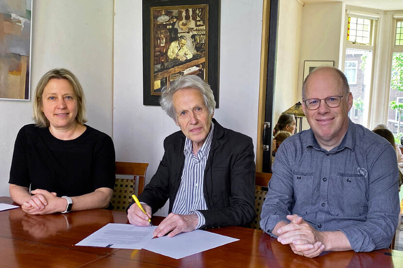 Drie mensen zitten aan een tafel in een woonkamer en kijken in de camera. De middelste persoon ondertekent een document.