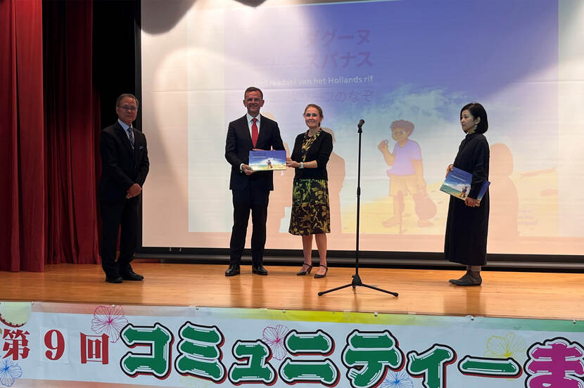 Links staat de heer Ikehiro Michio. In het midden staan Theo Peters en José Schreurs, waarbij José Schreurs het kinderboek aan Theo Peters overhandigd terwijl ze ook de hand schudden. Aan de rechterkant staat Kayoko Shimoji.