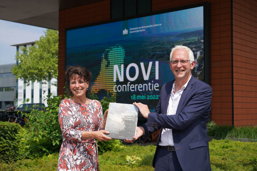 Twee mensen houden samen een boek vast en kijken glimlachend in de camera. Op de achtergrond is een banner te zien waarop staat 'NOVI conferentie, 18 mei 2022'.
