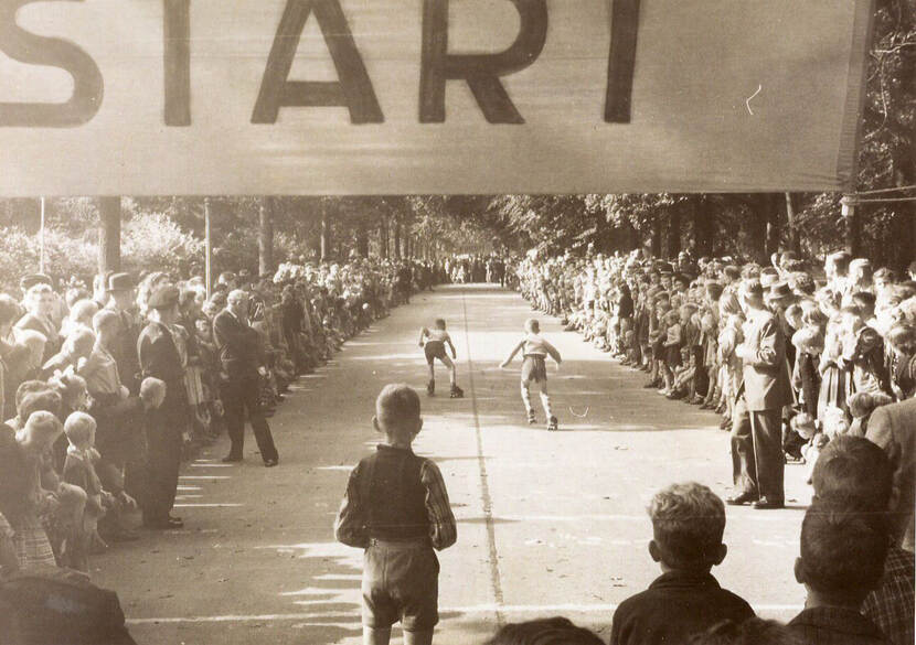 Zwartwit-foto van jongens die onder een banier door rolschaatsen waar 'Start' op staat. Langs de zijkanten van de weg staat publiek.