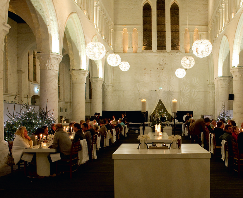 Mensen zitten aan gedekte tafels in een met kerstbomen en lichten versierde kerk.