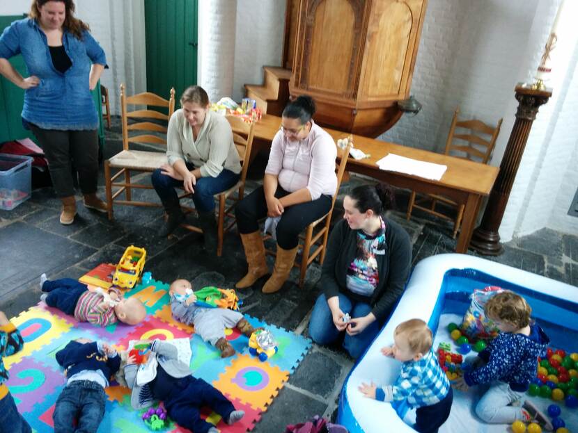 Moeders zitten op stoelen in een kerk en kijken naar baby's die op een matje op de grond liggen.