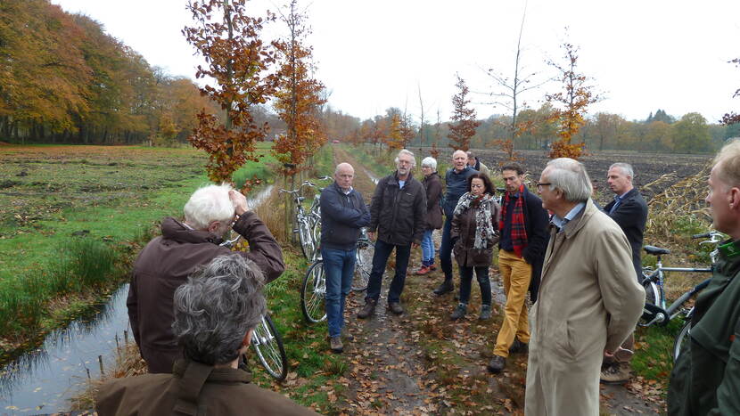 Een groep oudere mensen met fietsen staat op een pad waar jonge bomen staan.