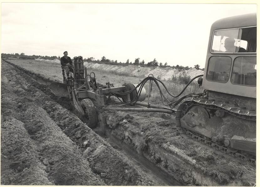 Zwart-wit foto van een man die met een ploeg achter een rupsbandwagen het land ploegt