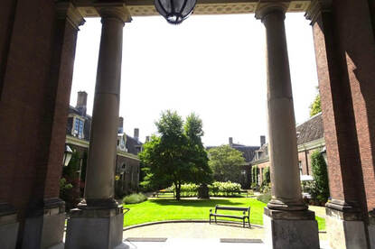 De binnenplaats van een kerk; gras en bomen met een bankje ervoor.