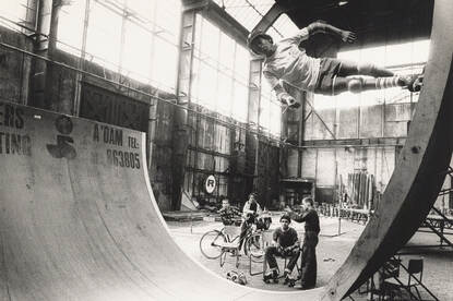 Zwart-wit foto van een jongen die op een halfpipe aan het skaten is, drie andere jongens zitten toe te kijken.